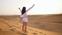 Young woman tourist in Dubai rear view from back stands in desert and stretches arms forward and sunbathes under sunlight enjoying warm weather. Traveling on vacation to beautiful places of world.