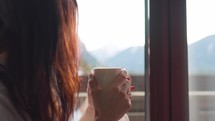 Young woman enjoying her morning coffee or tea, looking out the window at the snowy mountains. Vacation in the mountains. Great start to the day, time for new ideas.