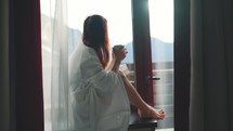 Young woman enjoying her morning coffee or tea, looking out the window at the snowy mountains. Vacation in the mountains. Great start to the day, time for new ideas.