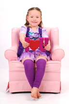 Young girl sitting in pink chair holding "I love you" sign.