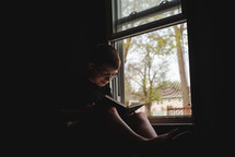 a boy reading a book in front of a window 