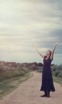 woman standing on a dirt road with hands raised praising God 