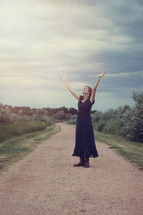 woman standing on a dirt road with raised arms in worship 