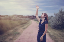 woman standing on dirt road with hand raised praising God 
