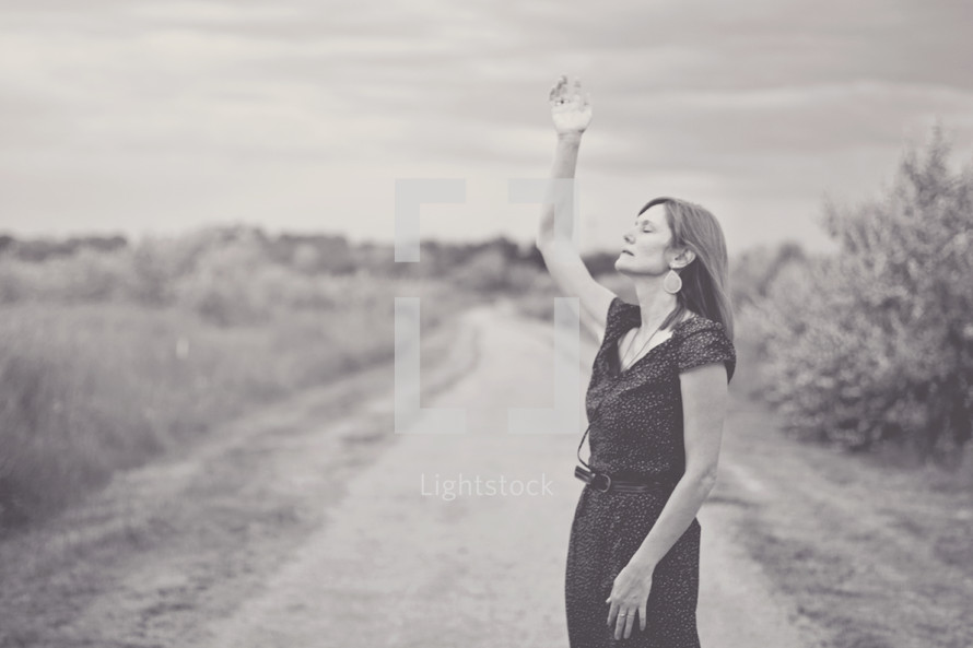 woman standing on a dirt road with a hand raised to praise God 