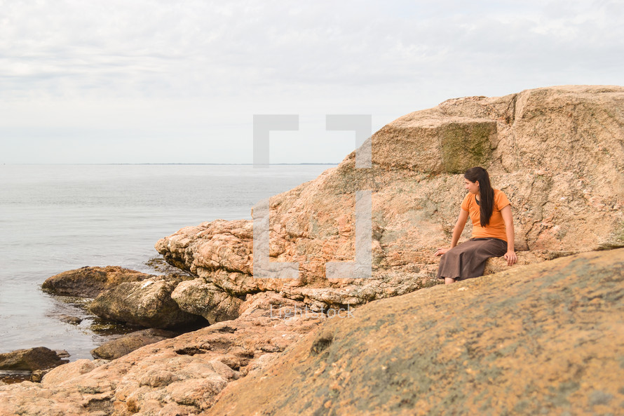 a woman sitting on a rock along a shore 