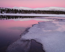 A tranquil winter scene reflecting in Grövelsjön lake, surrounded by snow-capped land and a pink sunrise sky.