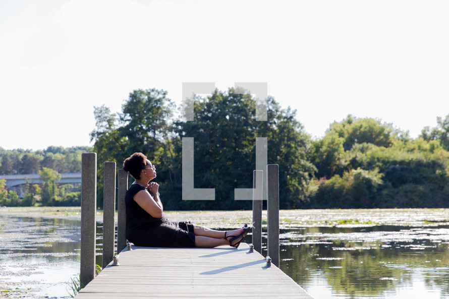 a woman with praying hands sitting at the end of a dock over a lake 