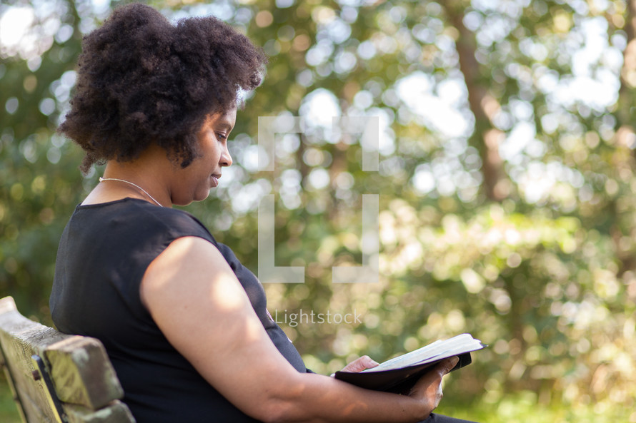an African American woman sitting on a bench reading a Bible outdoors 