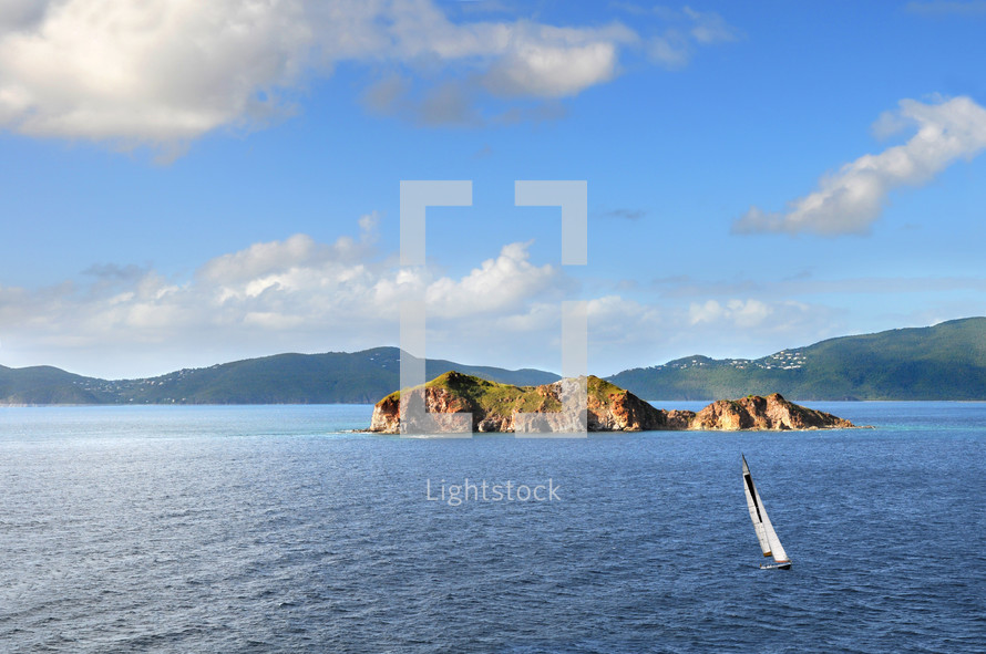 Rock islands in a body of water, a sailboat in the water and mountains in the background
