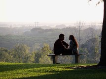 A young couple (man and woman) sit together on a park bench while the sun sets around then in  park with green grass.