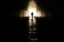 Man walking towards a light shaped cross formed by reflections on the walls, ceiling and floor of an underground concrete culvert.