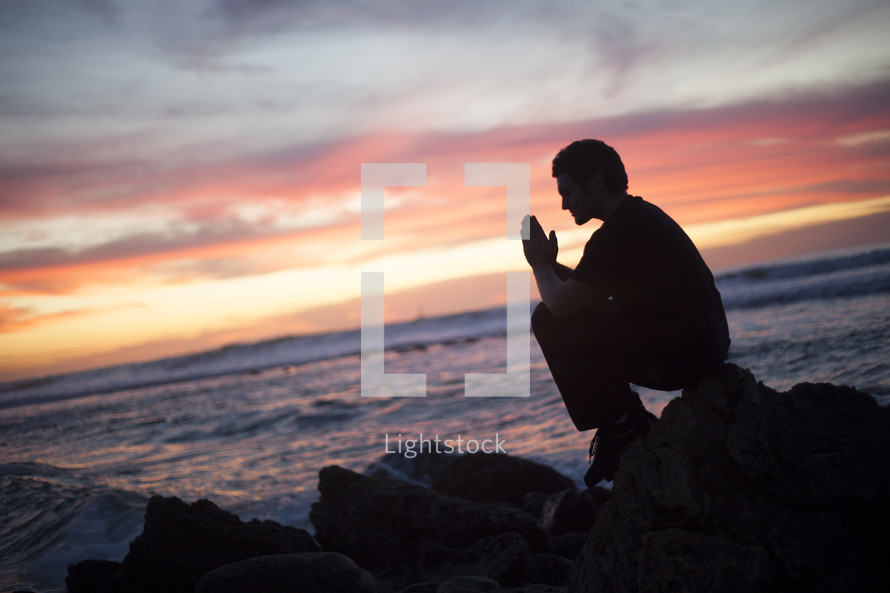 man in prayer sitting in prayer on a rock facing the ocean