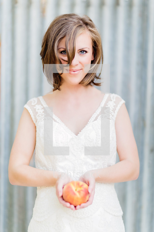 A woman holding a peach. Bride wedding white dress galvanized steel  Brunet brown hair 