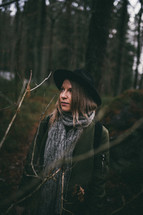 a woman in a coat, hat, and scarf standing in a forest 