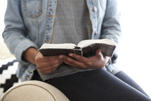 An African American woman sitting on the arm of a couch reading a Bible 