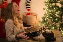 A laughing young woman sits by a Christmas tree holding a wrapped gift.