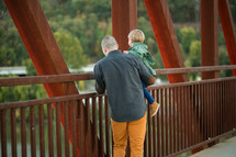 a father holding his son as they look over a bridge railing 