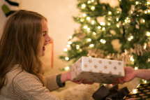 A laughing young woman near a Christmas tree gives a gift.