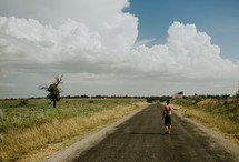 a boy child walking outdoors on a rural road carrying an American flag 