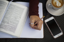 a young woman reading a Bible and writing in a journal at a table with a coffee mug and cellphone 