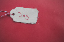 A Christmas gift tag reading "Joy," on a red background.
