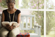 Smiling woman sitting on a porch swing outside, reading a Bible.