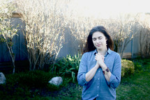 A woman standing outside in a garden praying with her eyes closed