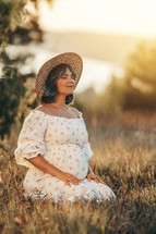 Happy Mother Carrying Child, Expecting For Baby. Amazing Motherhood. Pregnant Woman in beautiful dress and straw hat sitting on grass at nature rural meadow. High quality 