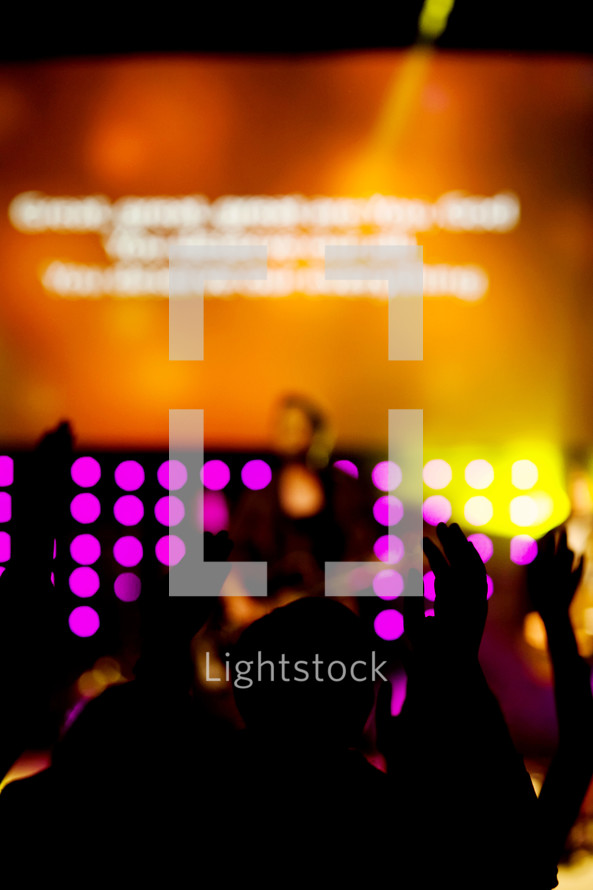 Silhouette of people with hands raised facing lighted stage during worship service