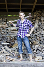 Man in rolled up denim jeans with axe over his shoulder standing in front of a wood pile.