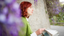 Beautiful red hair girl reading newspaper under wisteria purple flowers