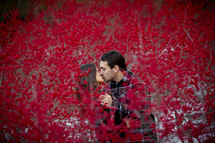 Couple kissing behind red flower blossoms.
