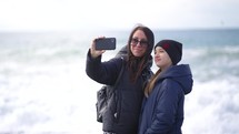 Cheerful young mother and daughter taking selfie on the beach. Smiling mother and daughter taking selfies at beach on a spring day. Family lifestyle concept.