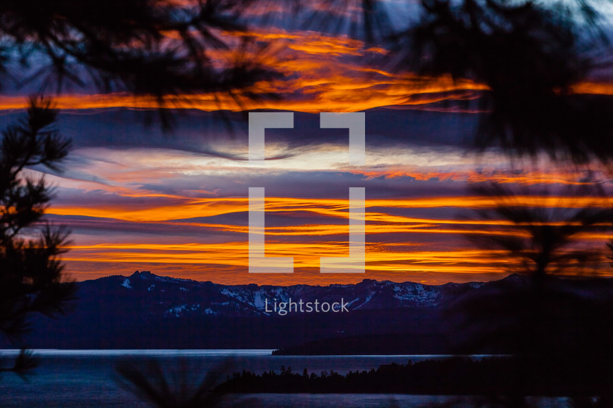 View thru pine trees of snow capped mountains lake & clouds at  orange sunset lake