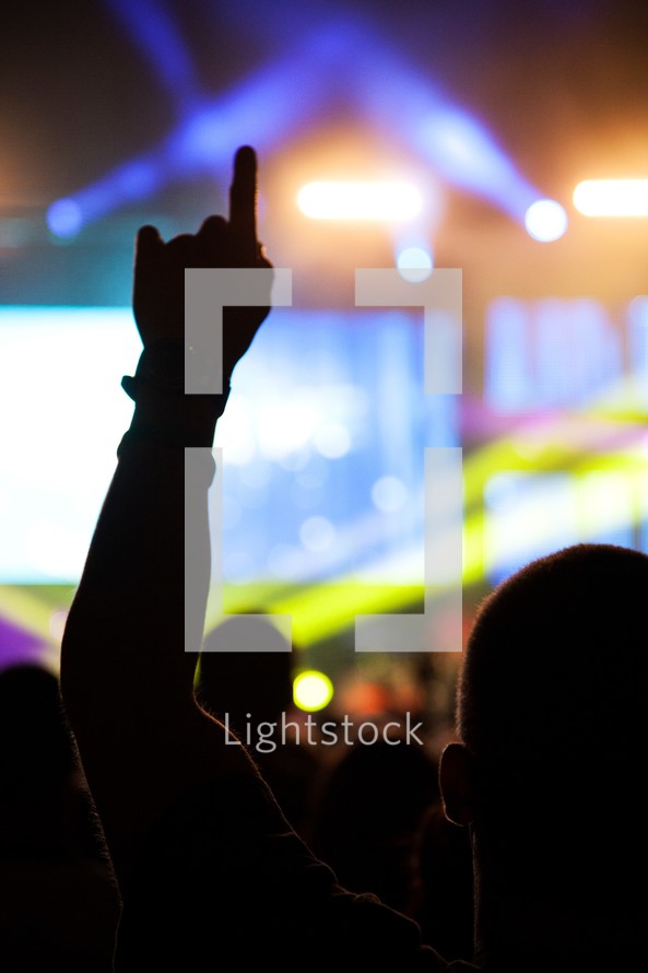 Silhouette of man watching concert on lighted stage with left hand pointer finger raised in the air.