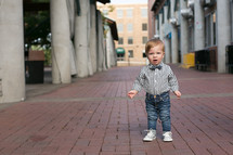 toddler boy in a bow tie standing on a sidewalk 