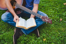 young man sitting in grass playing a guitar and reading a Bible 