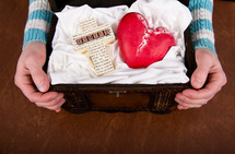 Hands holding a treasure chest containing a family cross and a red ceramic heart.