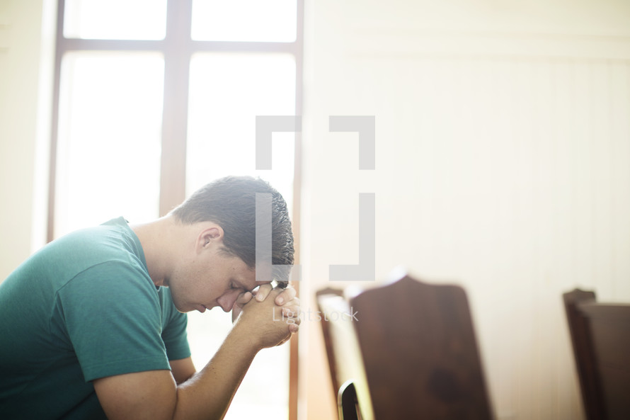Man with hands clasped and head bowed,praying in a church pew.