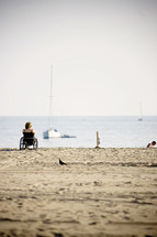 woman in a wheel chair on the sandy beach shore watching boats