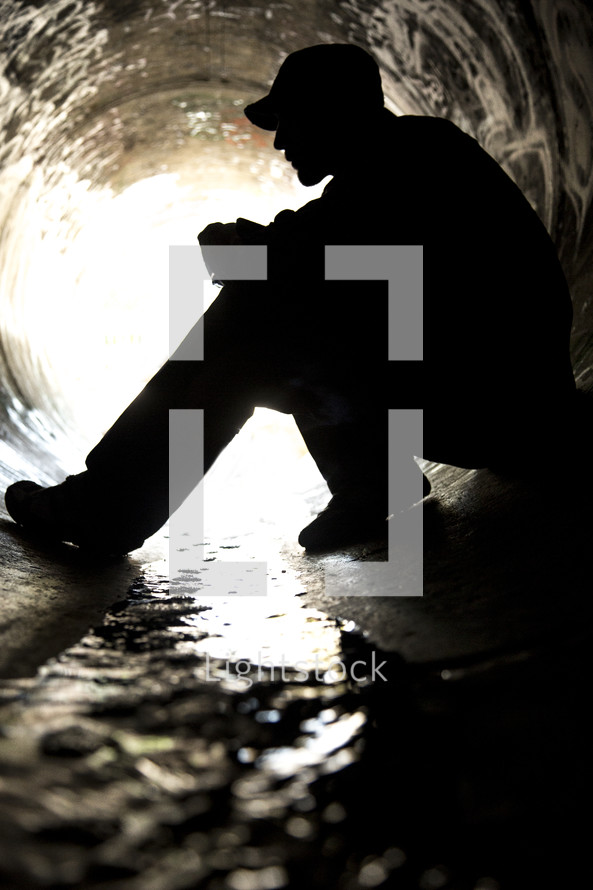 Silhouette of a man sitting in a sewer drian pipe.