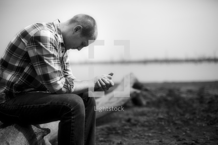 A man in a plaid shirt sitting on a log and praying.