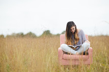 a young woman sitting in a chair in a field reading a Bible 