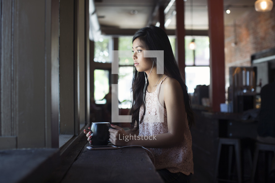 a young woman listening to earbuds in front of a window in a coffee shop 