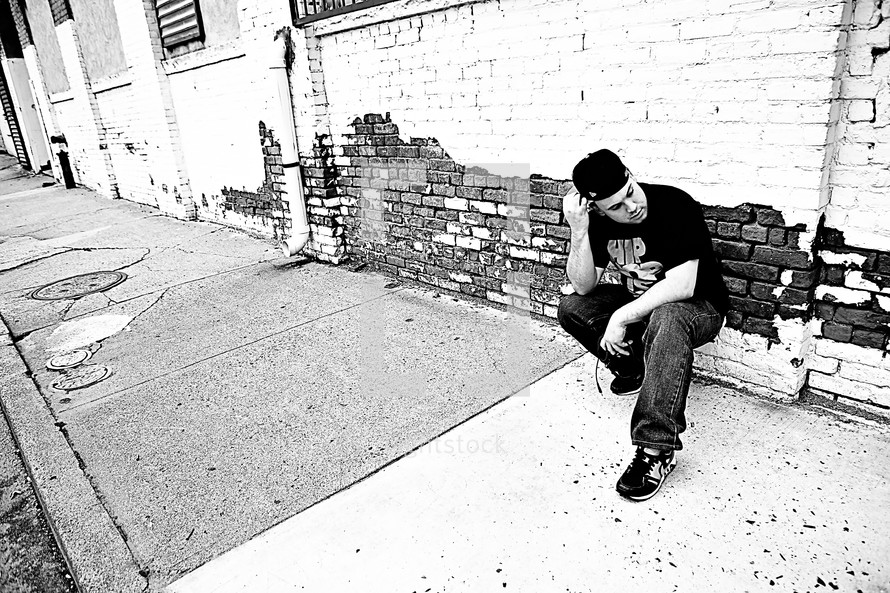 man crouched on a sidewalk in front of a brick wall