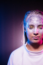 Woman standing in bright light with gold and silver makeup and glitter in her face. Colors in blue and pink wearing white clothing symbolizing an angel in modern shape