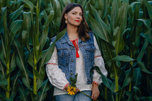 Smiling attractive hippy woman with sunflower on green corn nature background. Young lady in white embroidery shirt, denim waistcoat. Summer fashion, hipster, ethno, folk lifestyle.