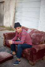 Young man in jeans with hat sitting on old sofa in alley outside garage door.