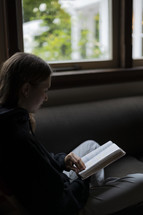 A young woman sitting in front of a large window reading her Bible